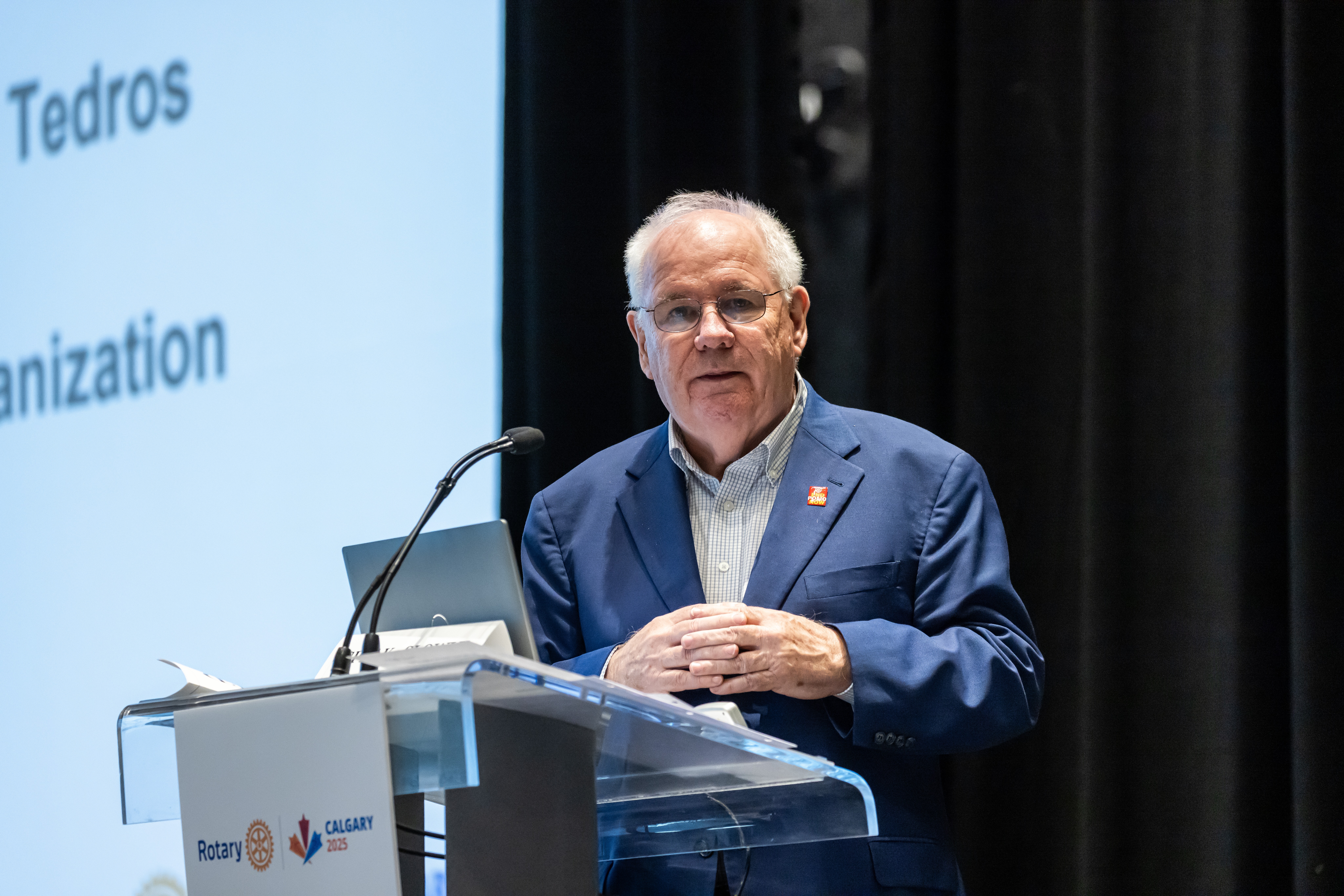O associado do Rotary Michael McGovern discursando em um pódio, com uma apresentação ao fundo. 