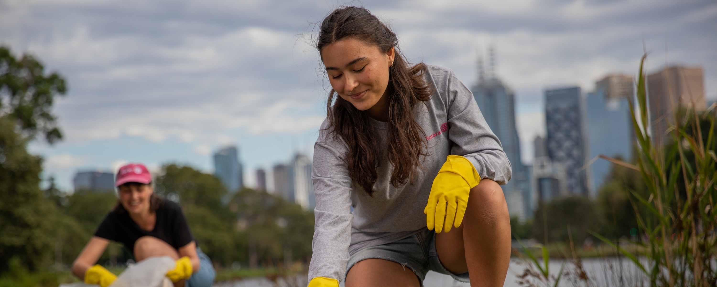 A woman wearing yellow gloves kneels outdoors and reaches toward the ground, with another volunteer and a city skyline behind her