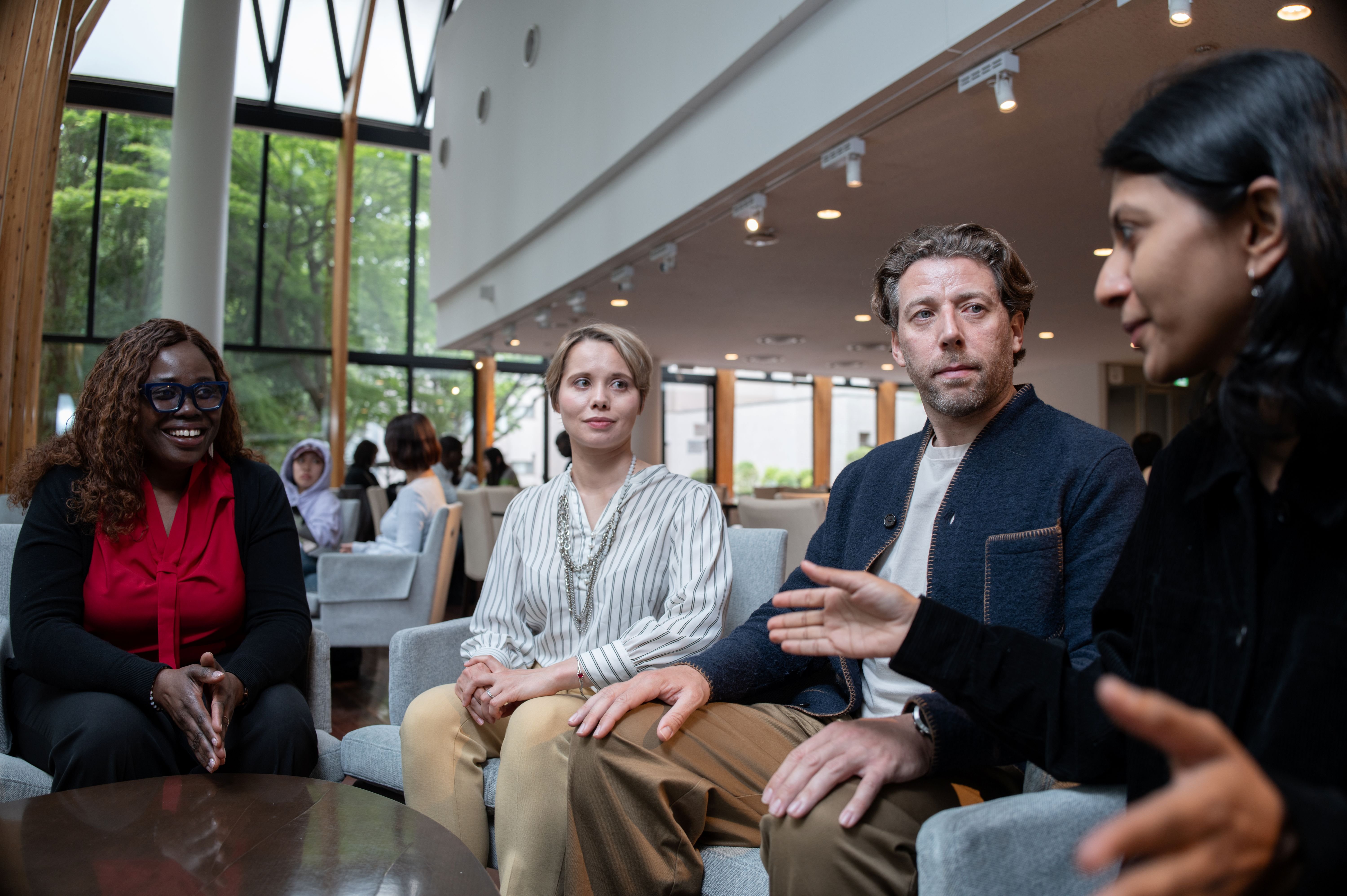 A group of people sit in a circle of chairs in a bright meeting space with large windows, listening and speaking during a small group discussion.