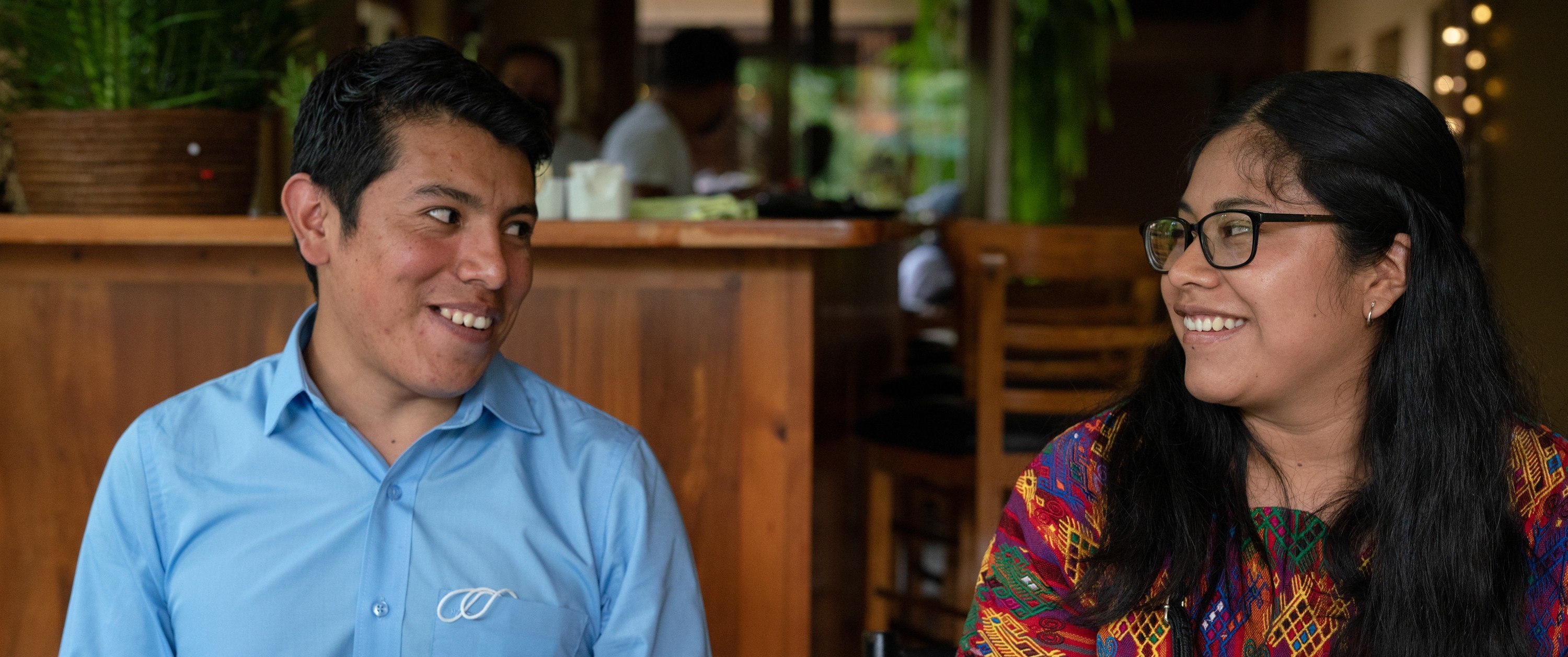Two people smile at each other while seated at a table in a café.