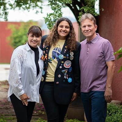 Three people standing together outdoors smiling, with trees and a brick wall in the background