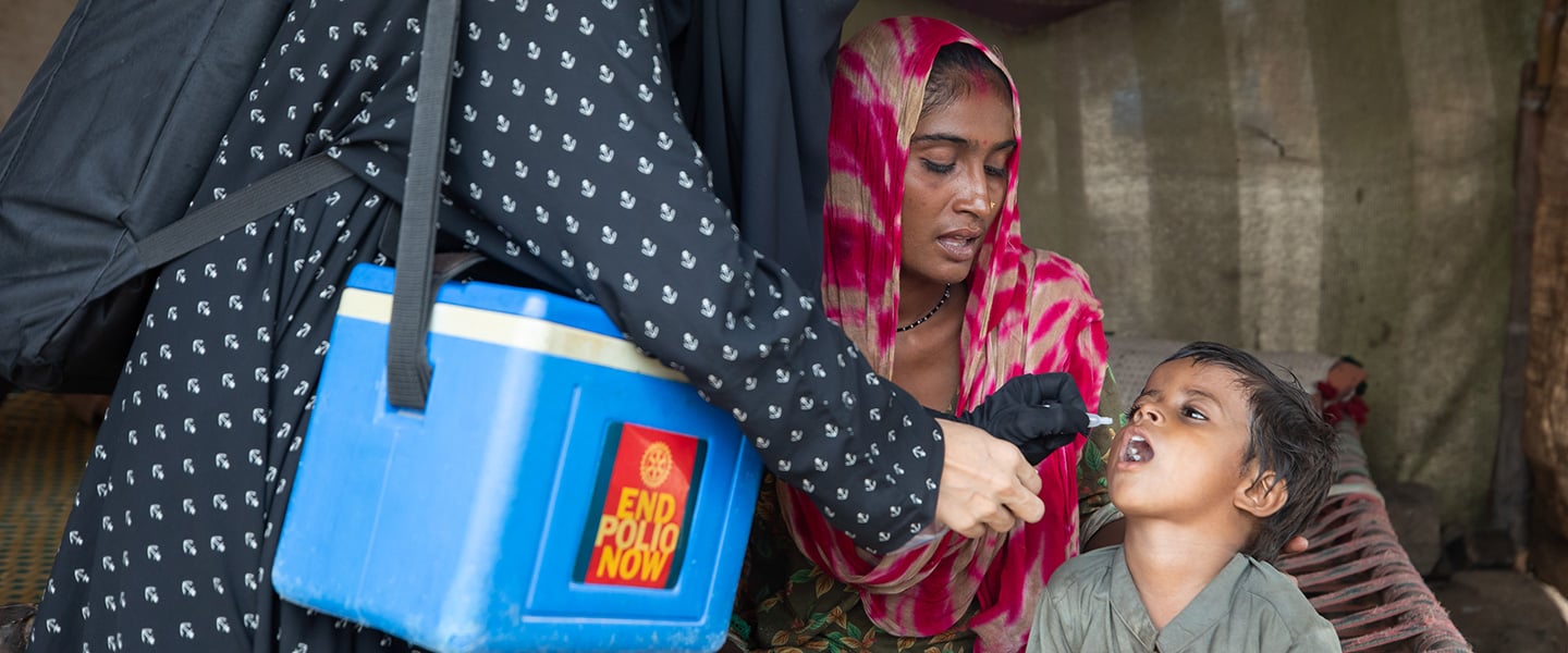 Health worker administers oral polio vaccine to a child during a door-to-door vaccination visit while a caregiver sits nearby.