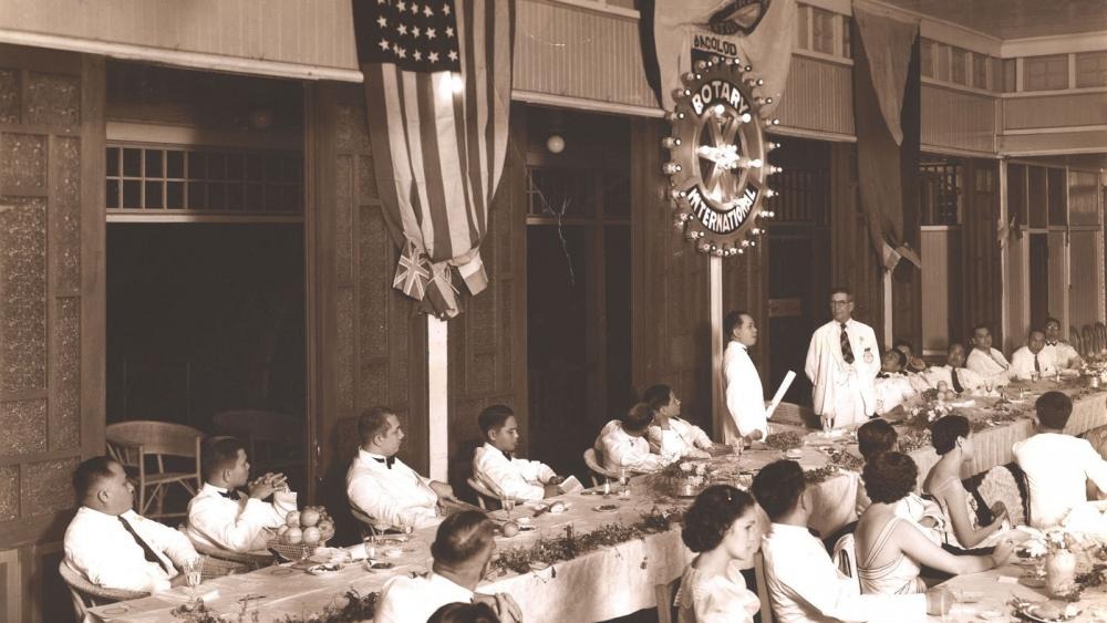 Historic photograph of a Rotary charter presentation ceremony, with attendees seated at long tables in a decorated hall beneath flags and a Rotary International sign.