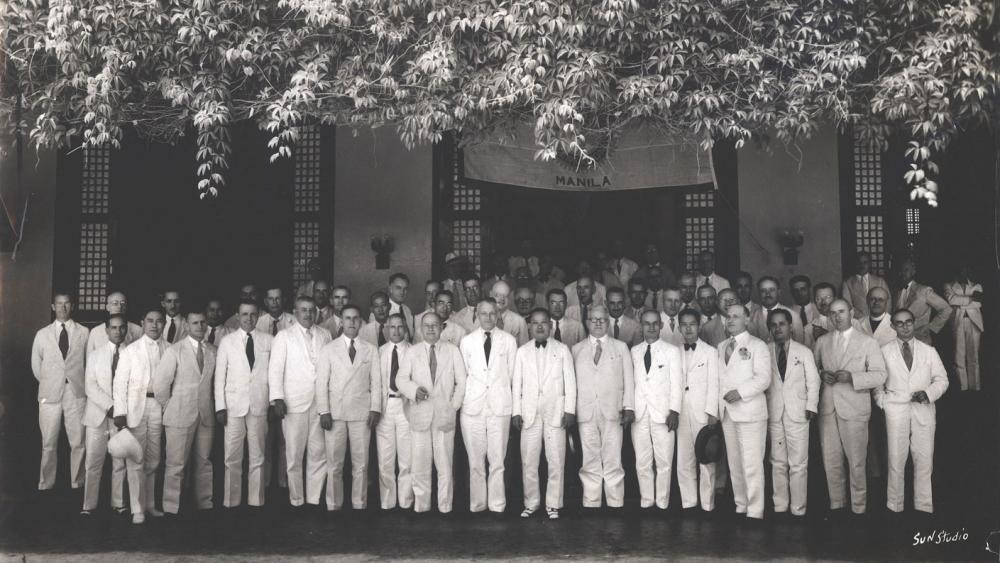 Historic group portrait of Rotary members standing together outside a building, with a banner overhead reading "Manila."