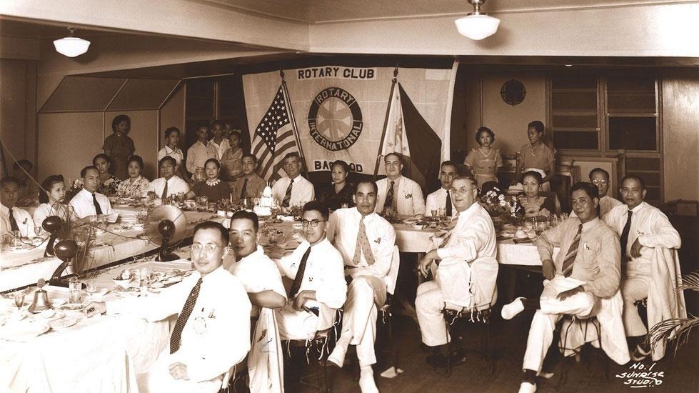Historic photograph of Rotary Club members and guests gathered around banquet tables with Rotary Club and Rotary International banners displayed in the background.