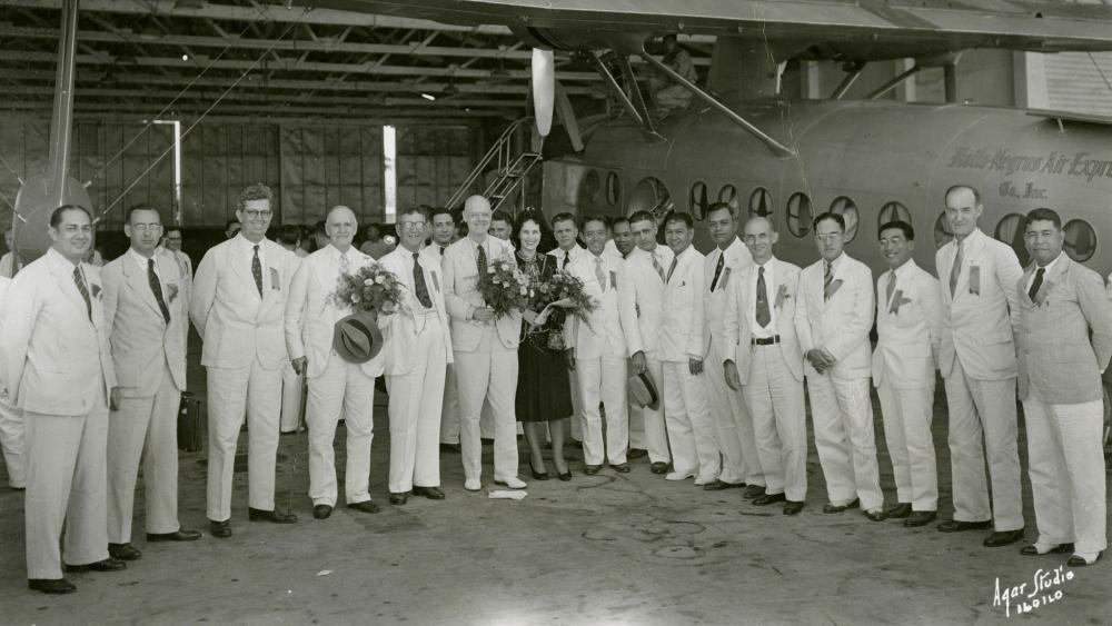 Historic photograph of Rotary members and guests standing in front of an airplane inside an aircraft hangar with flower bouquets presented to several participants.