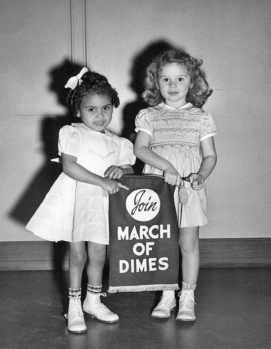 Schwarz-Weiß-Foto von zwei kleinen Kindern, die in einem Raum neben einer großen Sammelbox mit der Aufschrift „MARCH OF DIMES“ stehen. Ein Kind steht hinter der Kiste, während das andere daneben steht. Beide Kinder tragen Kleider und halten Puppen in den Händen. Das Foto wurde vor einem schlichten Hintergrund aus Wand und Boden inszeniert.