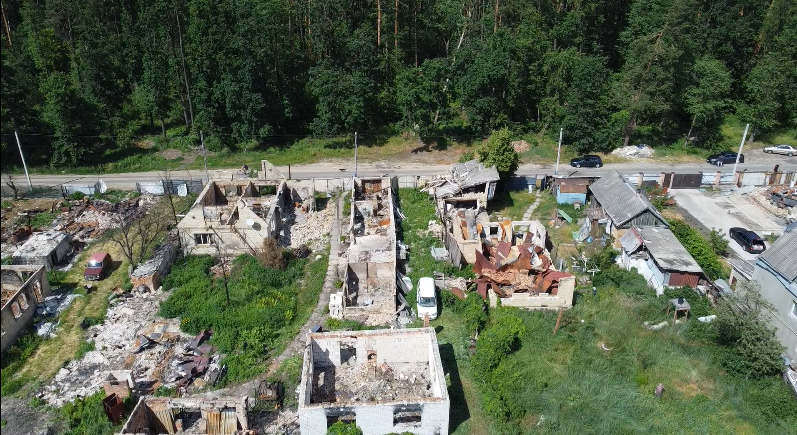 災害により複数の建物が被害を受けたウクライナの住宅街の空撮写真です。屋根が崩れ落ち、内部がむき出しになり、周辺一帯に瓦礫が散乱している様子が写っています。いくつかの建物は一部がまだ残っていますが、他の建物は甚大な被害を受けています。被災した地域社会は木々に囲まれており、画面の端には道路や近隣の建物が見えます。