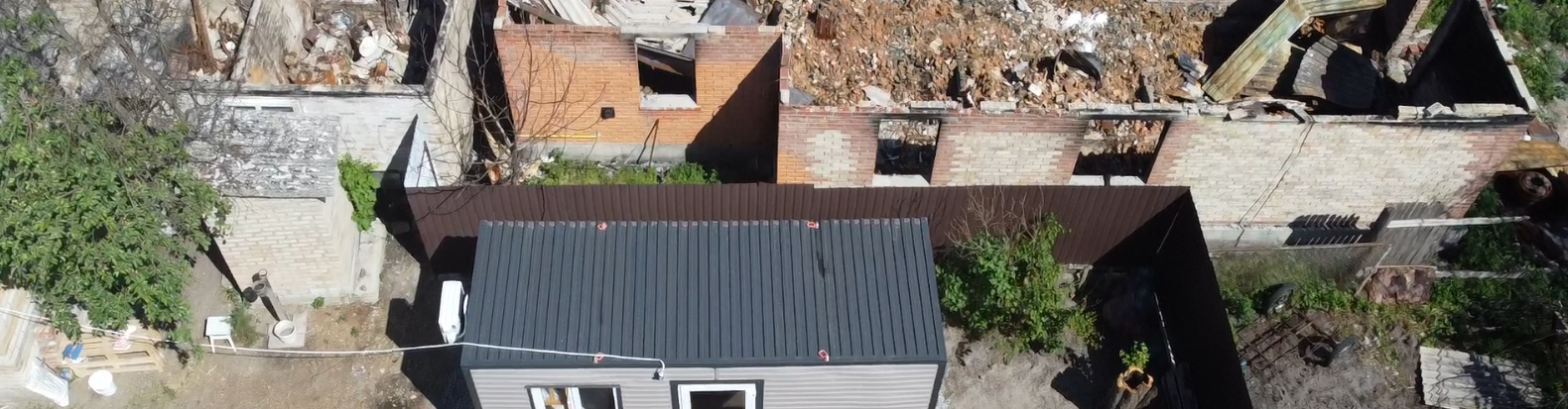 災害により複数の建物が被害を受けたウクライナの住宅街の空撮写真です。屋根が崩れ落ち、内部がむき出しになり、周辺一帯に瓦礫が散乱している様子が写っています。いくつかの建物は一部がまだ残っていますが、他の建物は甚大な被害を受けています。被災した地域社会は木々に囲まれており、画面の端には道路や近隣の建物が見えます。