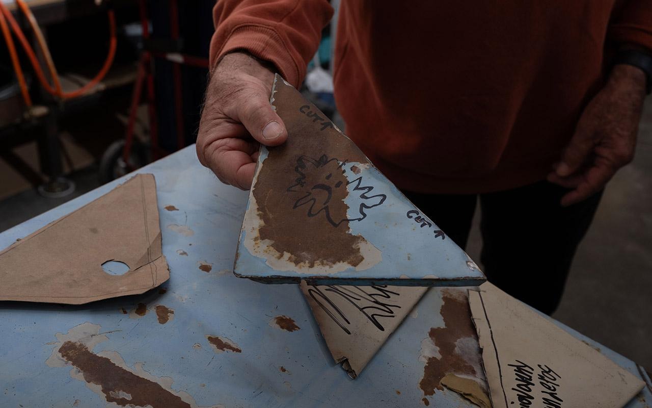 Uma pessoa segura um gabarito metálico triangular com marcações feitas a caneta sobre uma mesa de trabalho onde há outros gabaritos.