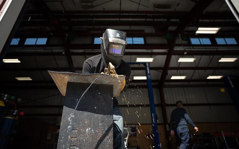 Um aluno solda um painel de metal usando um capacete de soldagem em uma oficina.