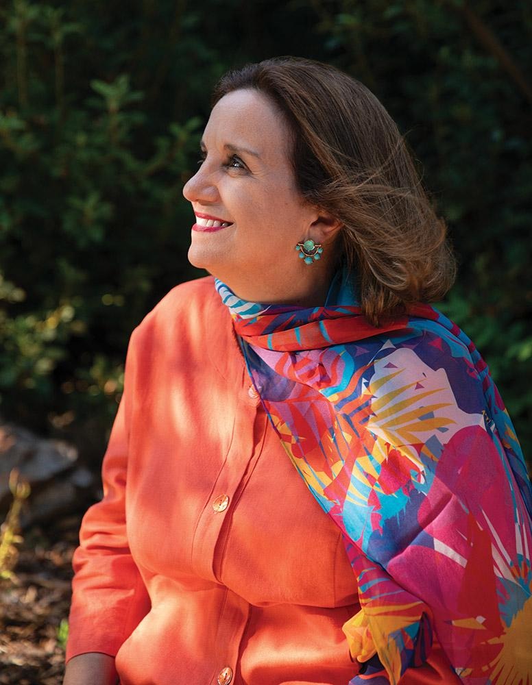 Isabel Scarinci seated outdoors in dappled sunlight, wearing an orange jacket and a colorful patterned scarf.