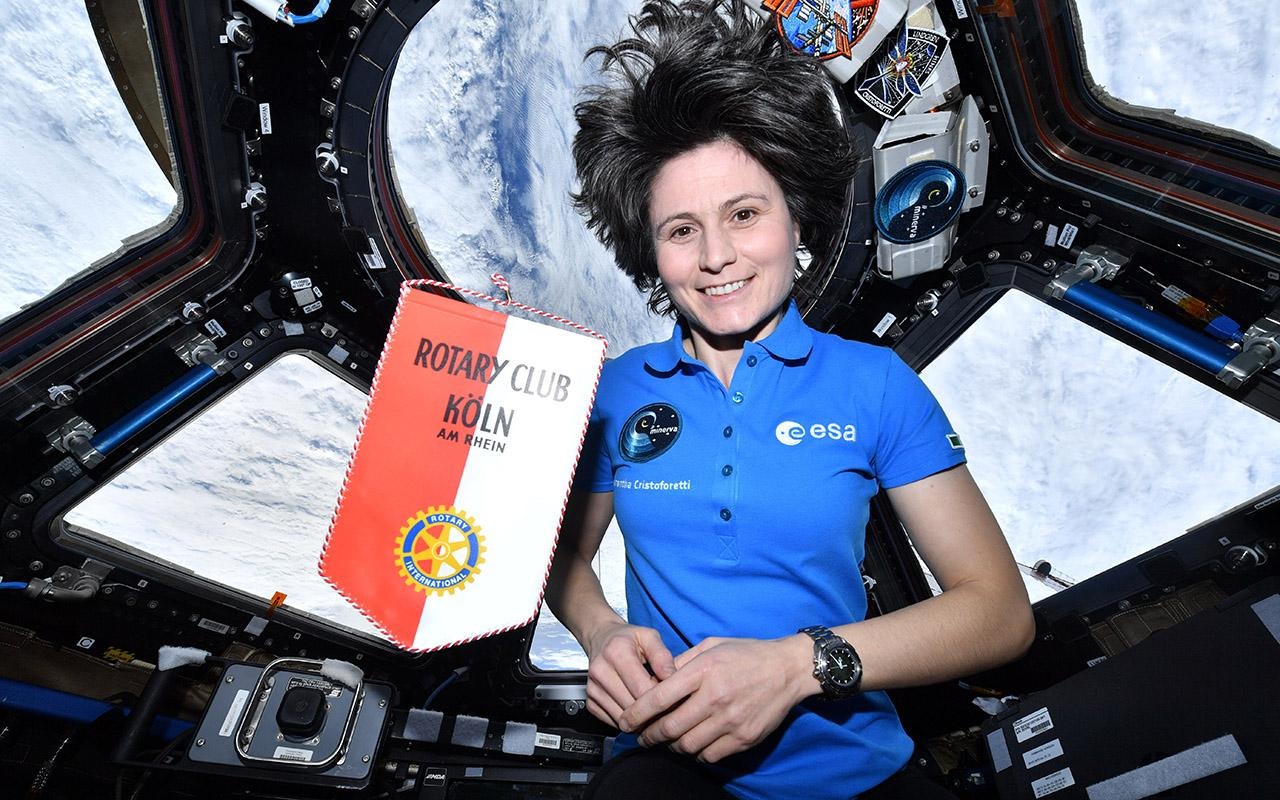 Samantha Cristoforetti sostiene un folleto del Plan de Acción de Rotary dentro de una nave espacial, desde cuya ventana se ve la Tierra.