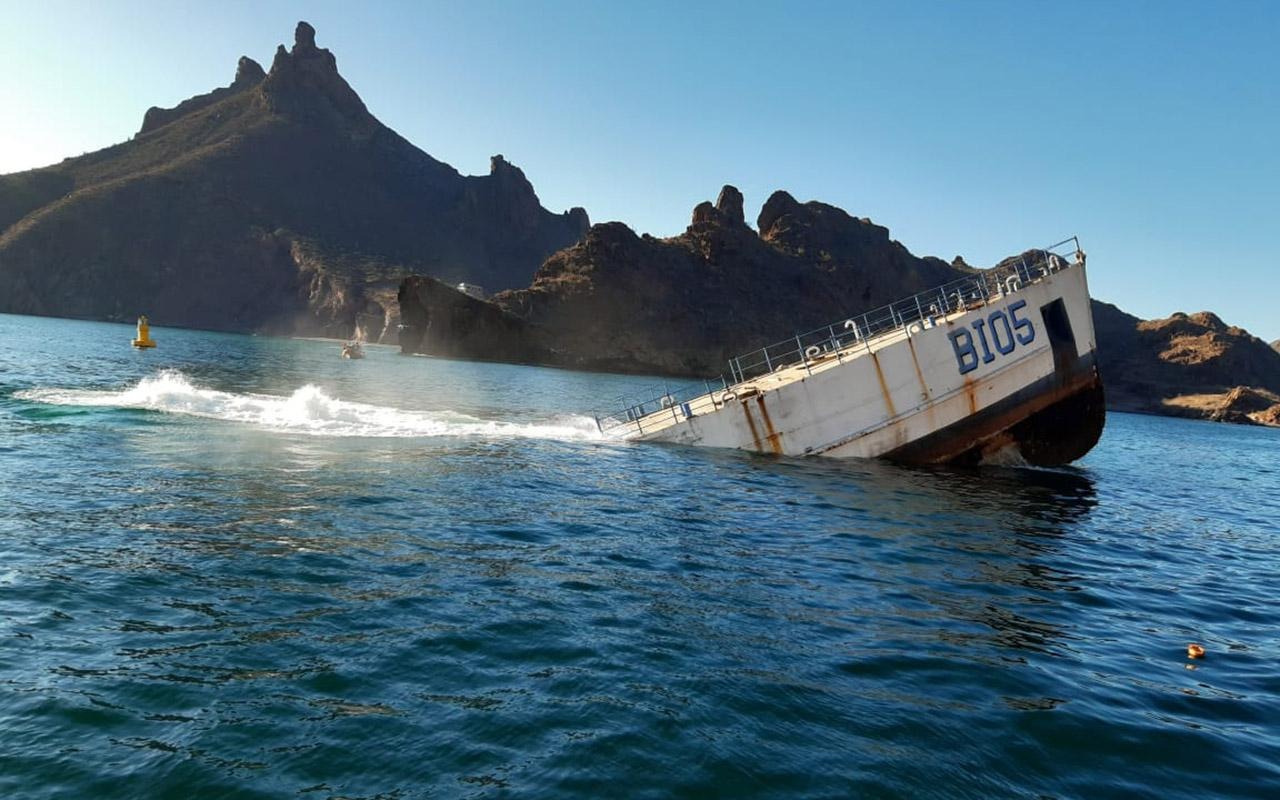 A sinking 190-foot boat in coastal water with mountains in the background and "blue" visible on the hull.