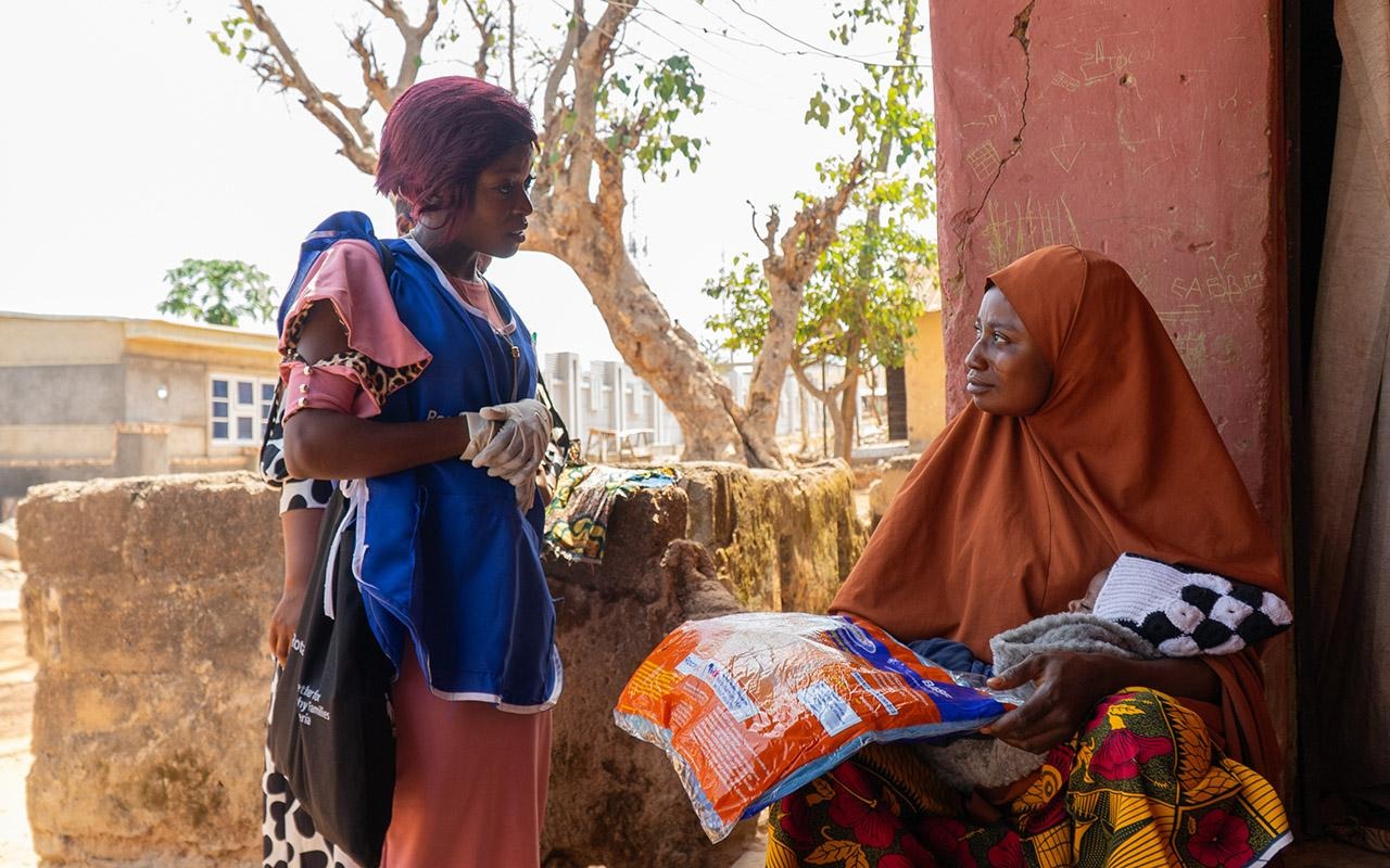 Eine lokale/r Gesundheitshelfer/in in Nigeria, der/die eine Rotary-Freiwilligenweste trägt, unterhält sich vor einem Gebäude mit einer sitzenden Person, die ein Paket in der Hand hält.