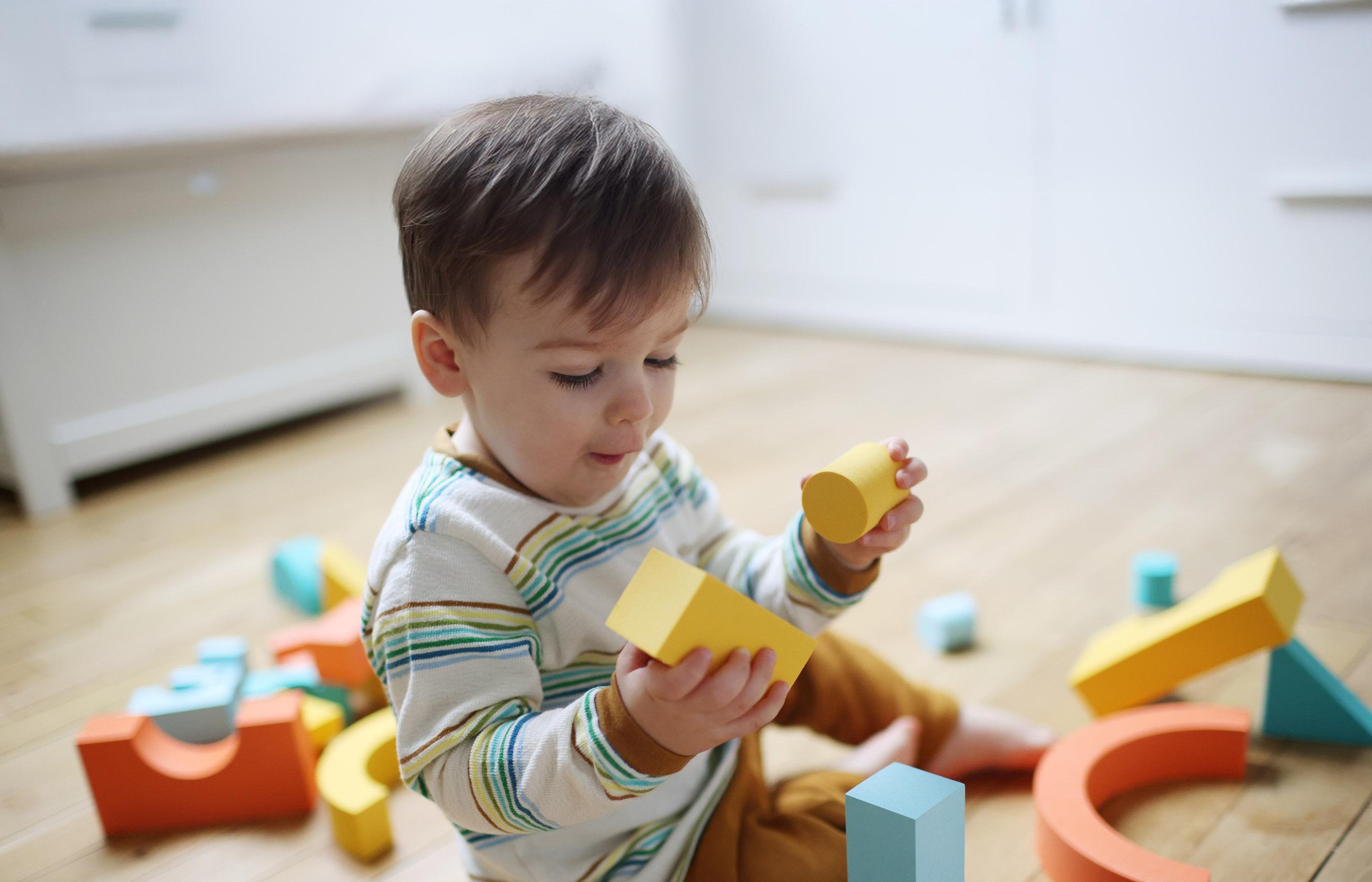 Ein Kind sitzt auf einem Holzboden, hält gelbe Bausteine in den Händen und spielt mit bunten geometrischen Formen.
