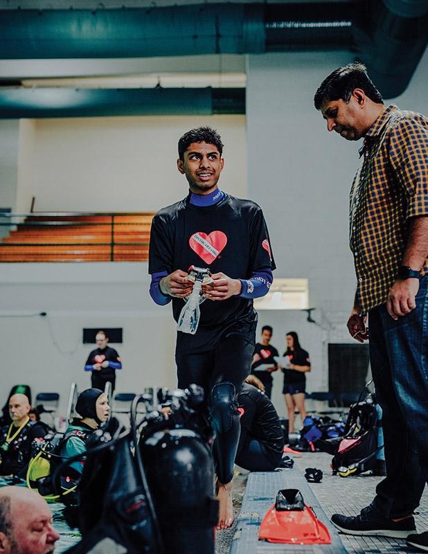 Adaptive diver Adarsh Akula in a wetsuit walking beside scuba tanks and diving equipment at an indoor pool facility.