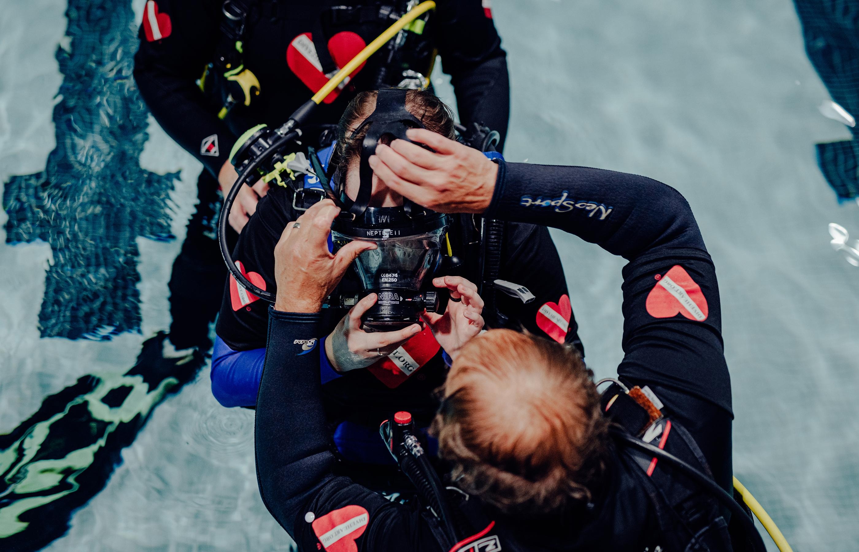 A scuba instructor puts on a diver mask on a student in a poo
