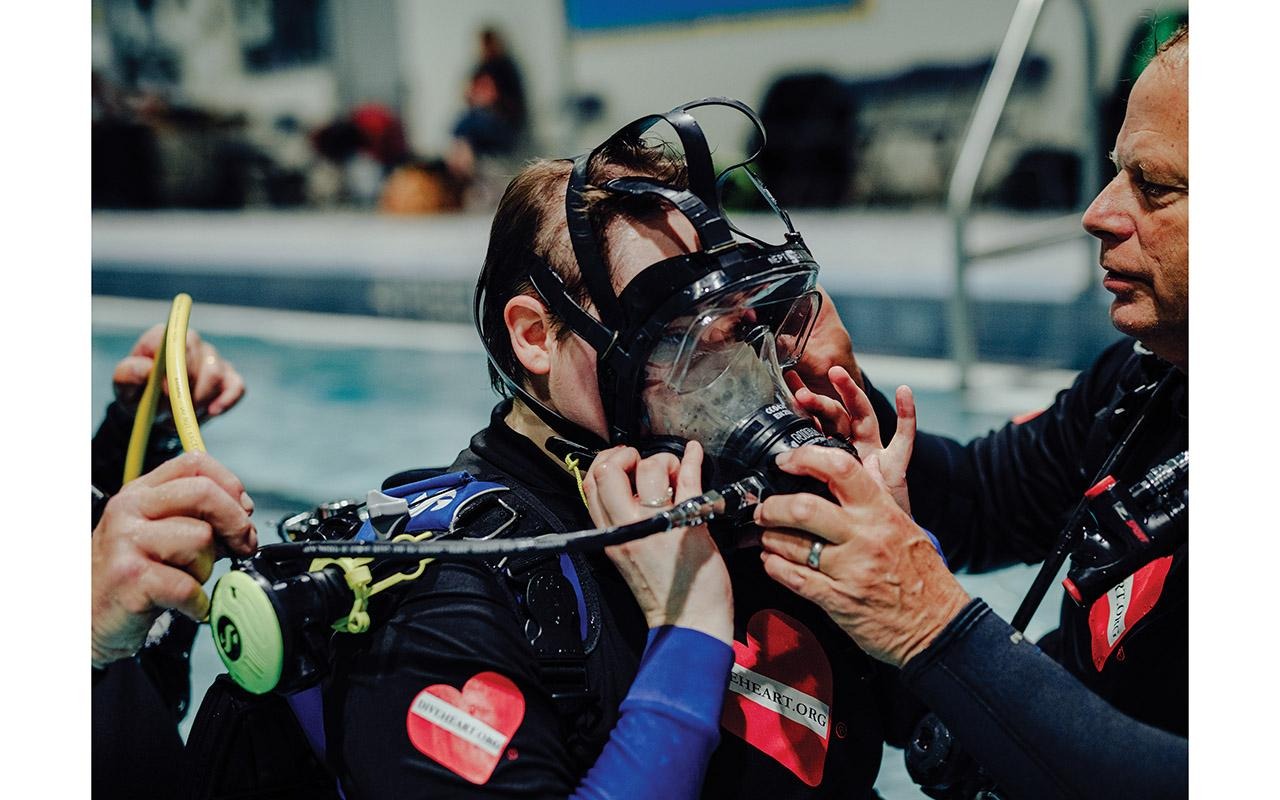 Scuba instructor helping another person adjust a full-face diving mask beside an indoor pool.