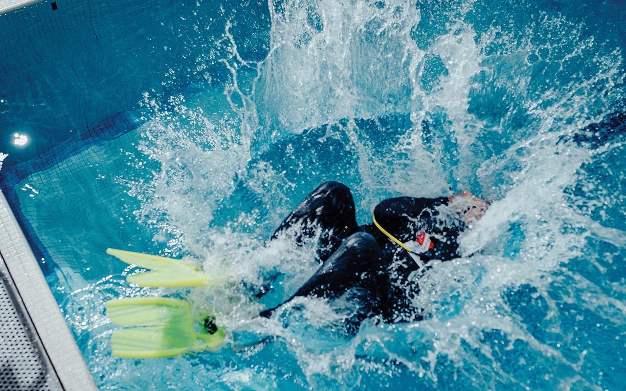 Person entering a pool from the edge while wearing scuba gear and fins as water splashes around them.