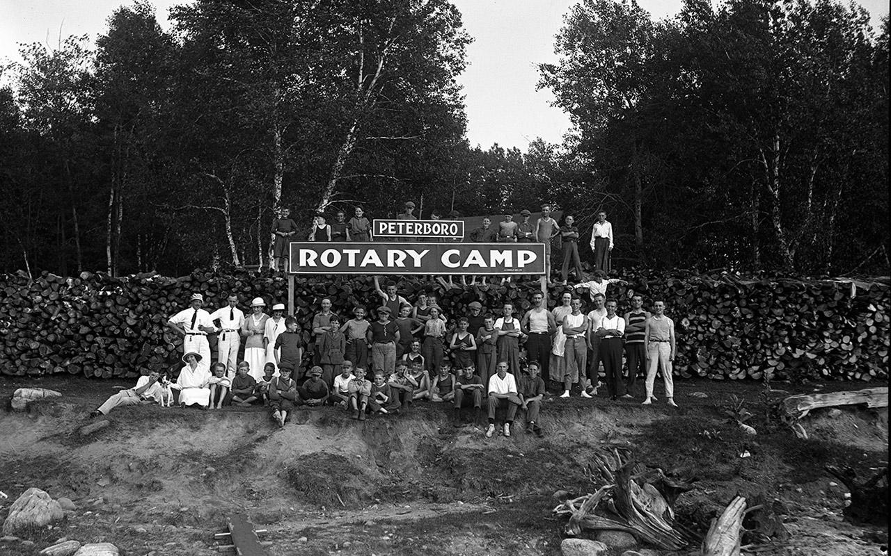 Group of people gathered in front of a sign reading "ROTARY CAMP" in a black-and-white outdoor photograph.