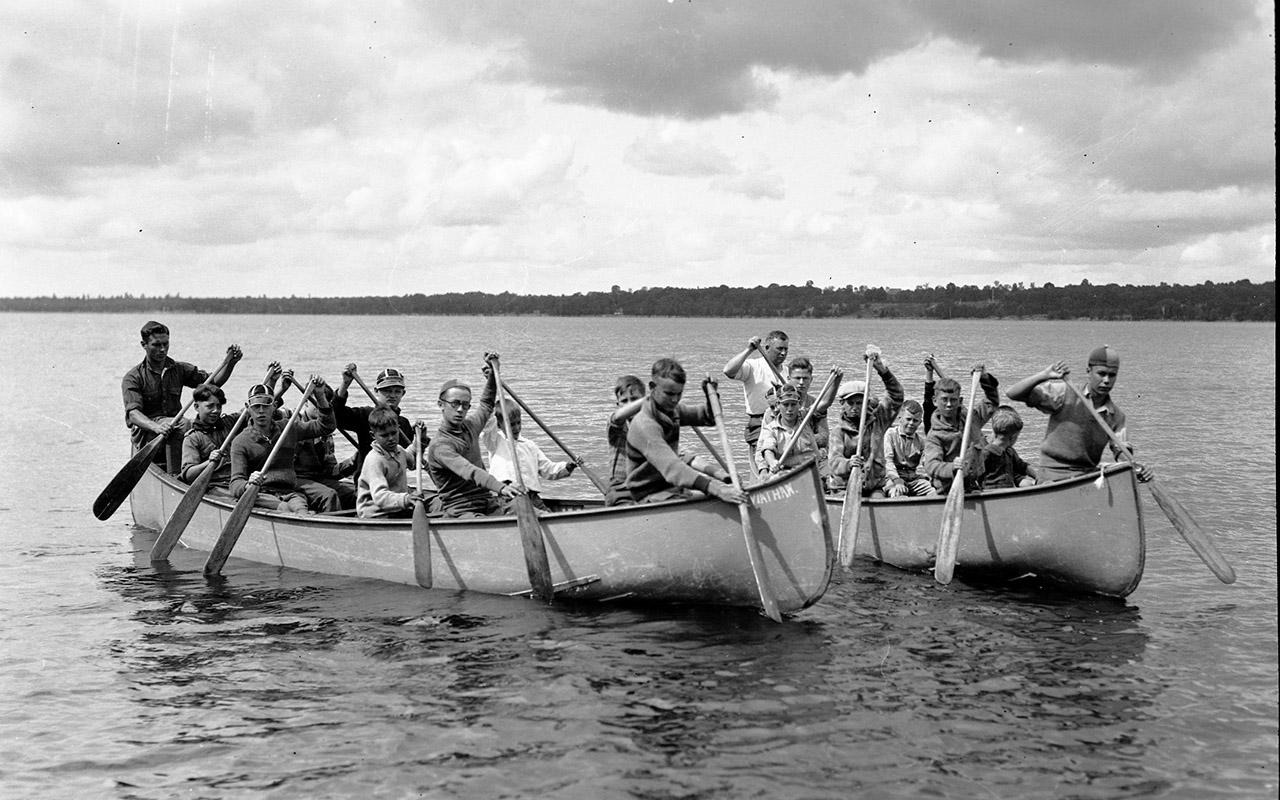 Campers paddling two canoes across a lake in a black-and-white photograph.
