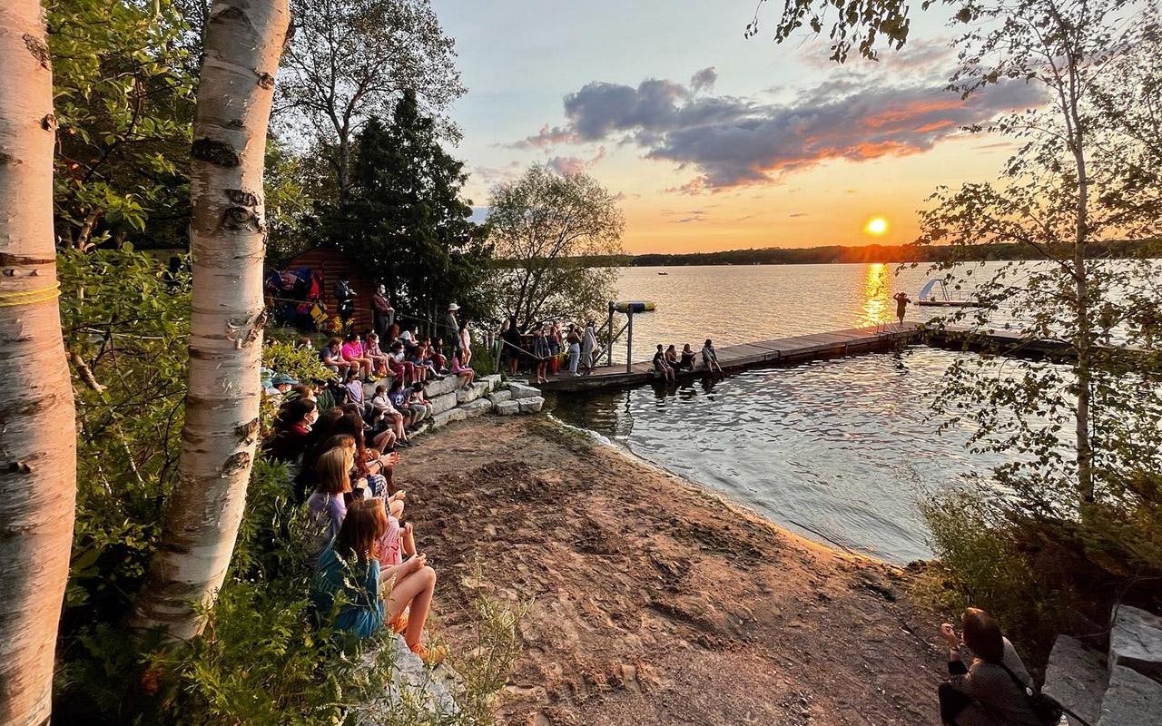 Campers seated along a lakeshore and dock at sunset beside a lake surrounded by trees at Camp Kawartha.