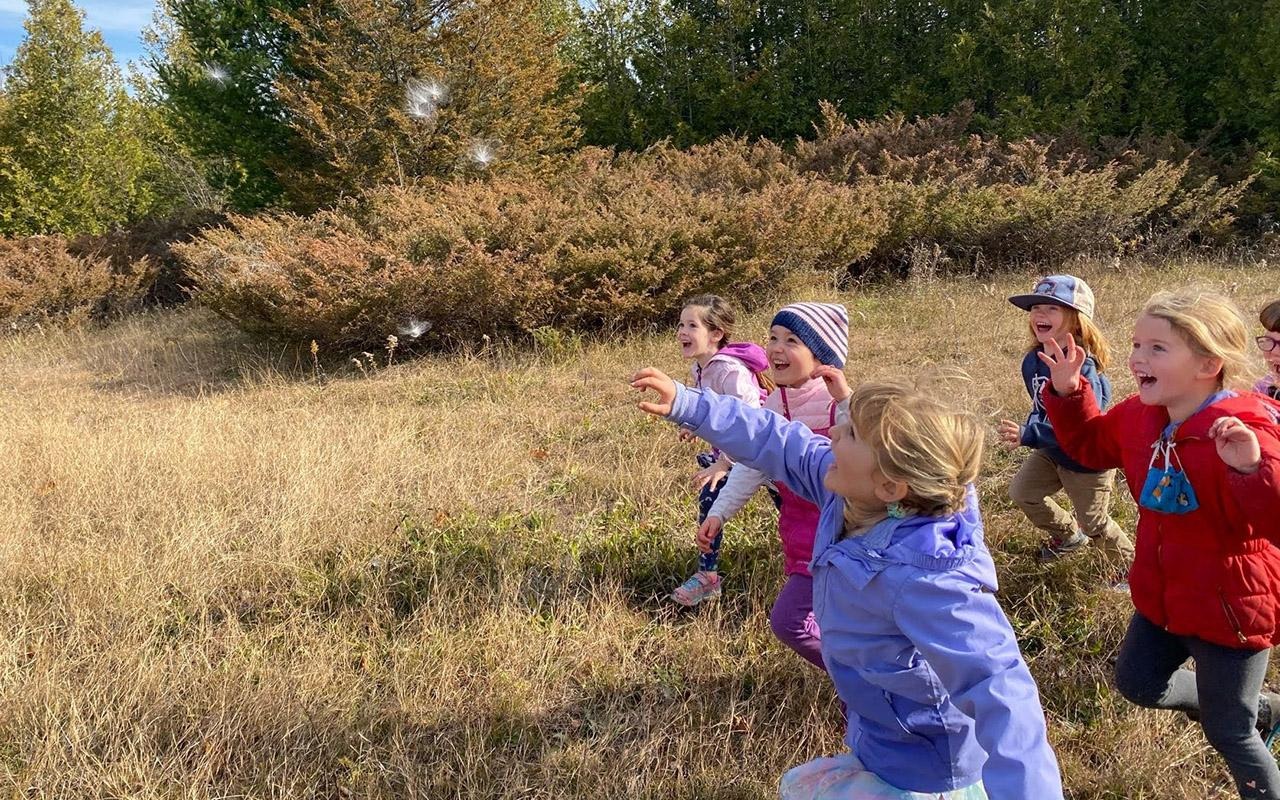 Campers in colorful jackets pointing and gesturing in a grassy field with trees in the background.