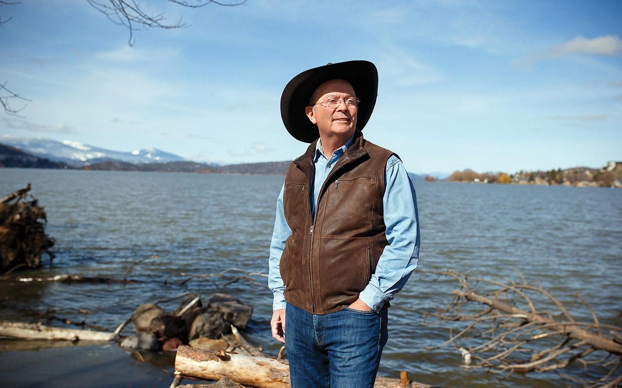 Rotary member Jim Root standing on a rocky lakeshore with hands in pockets, wearing a hat and looking across the water.