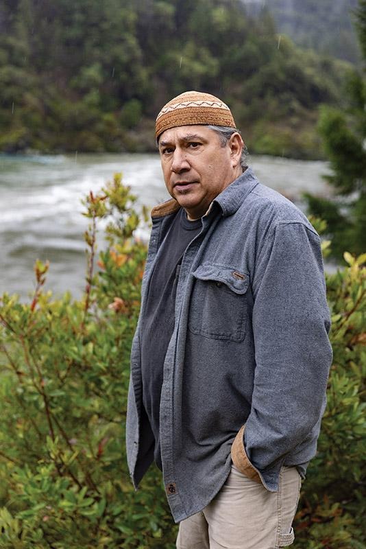 Leaf Hillman standing beside shrubs near a river, with forested hills and water in the background.