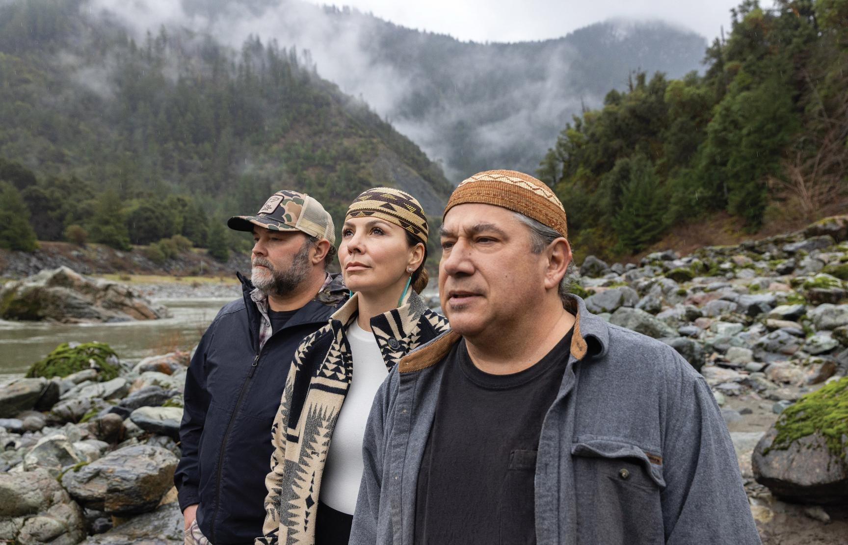 Three people standing on a rocky riverbank with forested mountains and low clouds in the background.