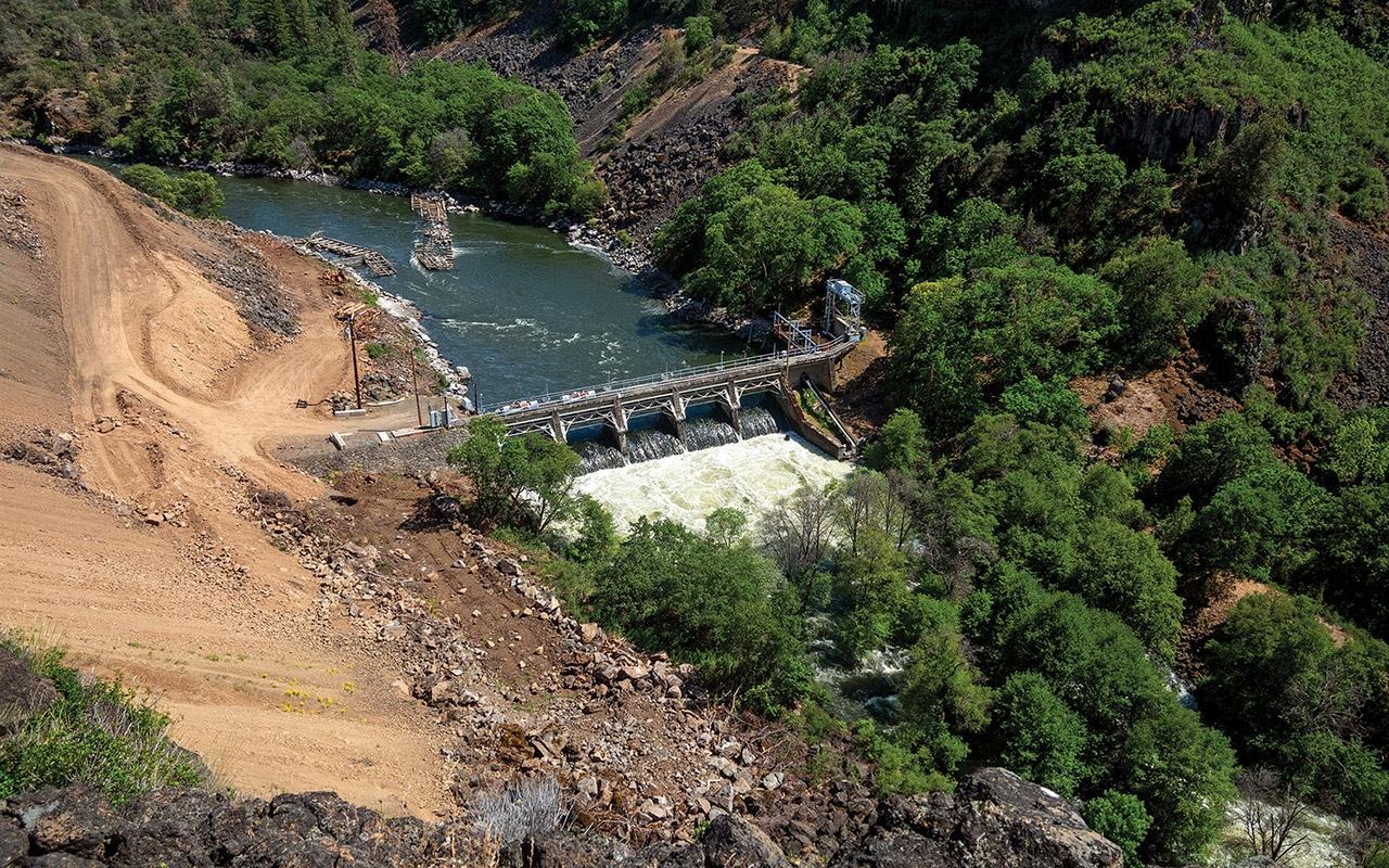 Aerial view of a river, damaged roadway, and bridge surrounded by forested hills.