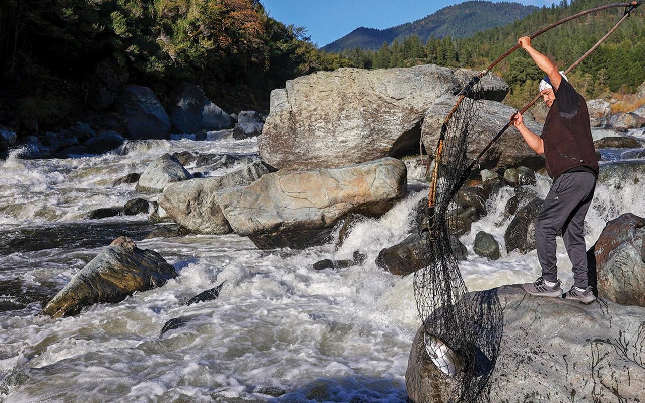 A Karuk tribal member standing on riverside rocks and lifting a fish net above fast-moving water.