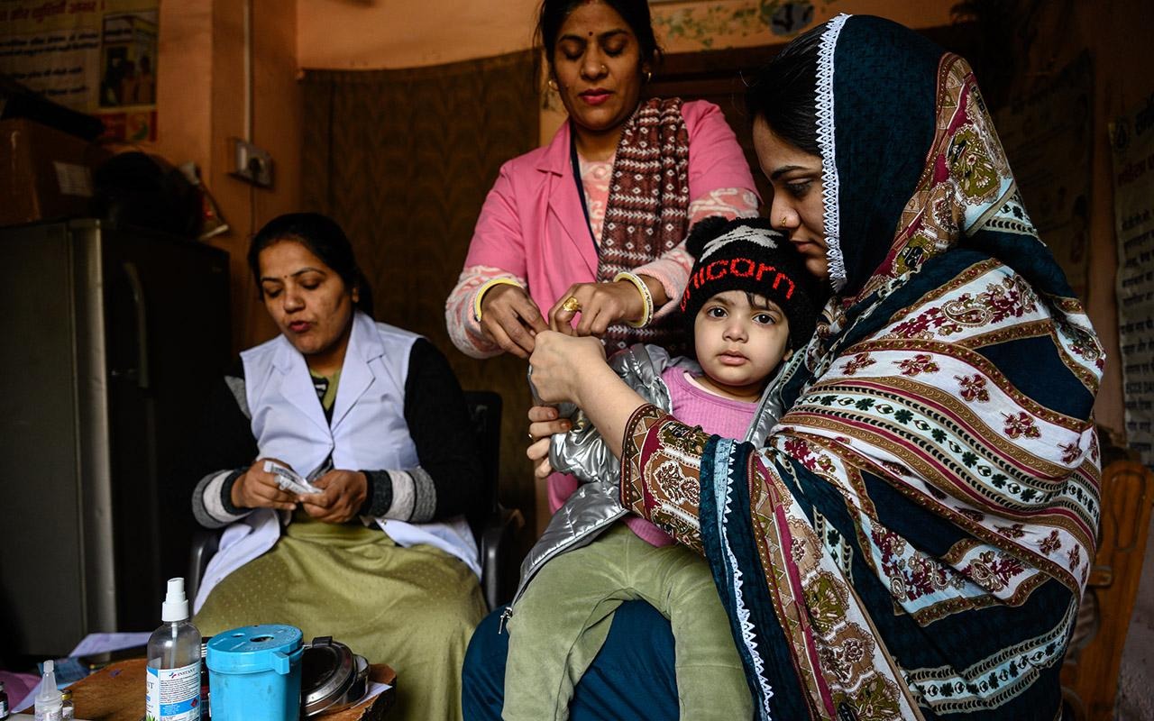 Gli operatori sanitari comunitari che assistono un bambino mentre un’altra persona prepara il materiale in un ambulatorio al chiuso.