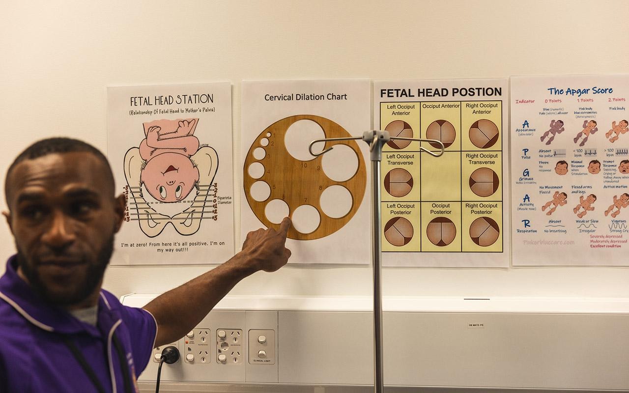 Simon James Kopalua désigne un tableau de dilatation cervicale situé à côté d'affiches intitulées « Position de la tête du fœtus », « Orientation de la tête du fœtus » et « Score d'Apgar »