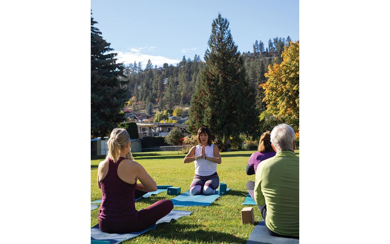 People seated on yoga mats outdoors while an instructor leads a group session in a park surrounded by trees