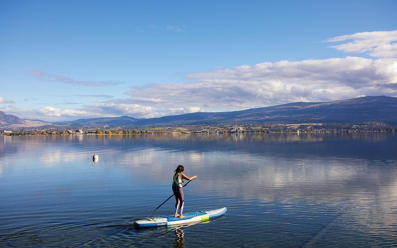 Julie Mulligan standing on a paddleboard on a calm lake with mountains and shoreline buildings in the background