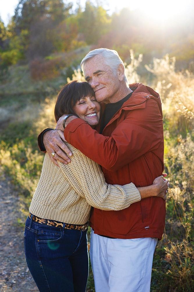 Julie Mulligan and her husband John embracing on a sunlit path surrounded by tall grass and trees outdoors