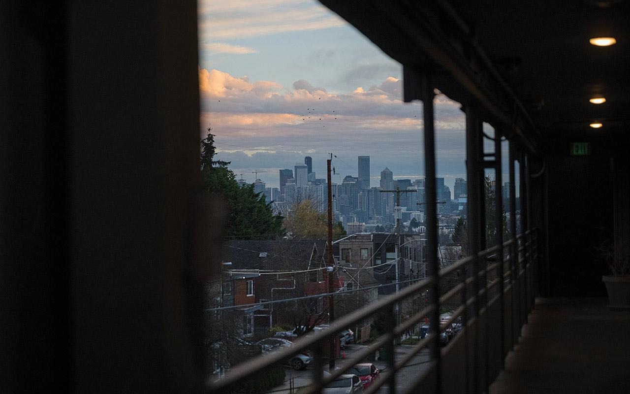 Exterior corridor with metal railing overlooking Seattle skyline at dusk, framed by building columns and lights overhead