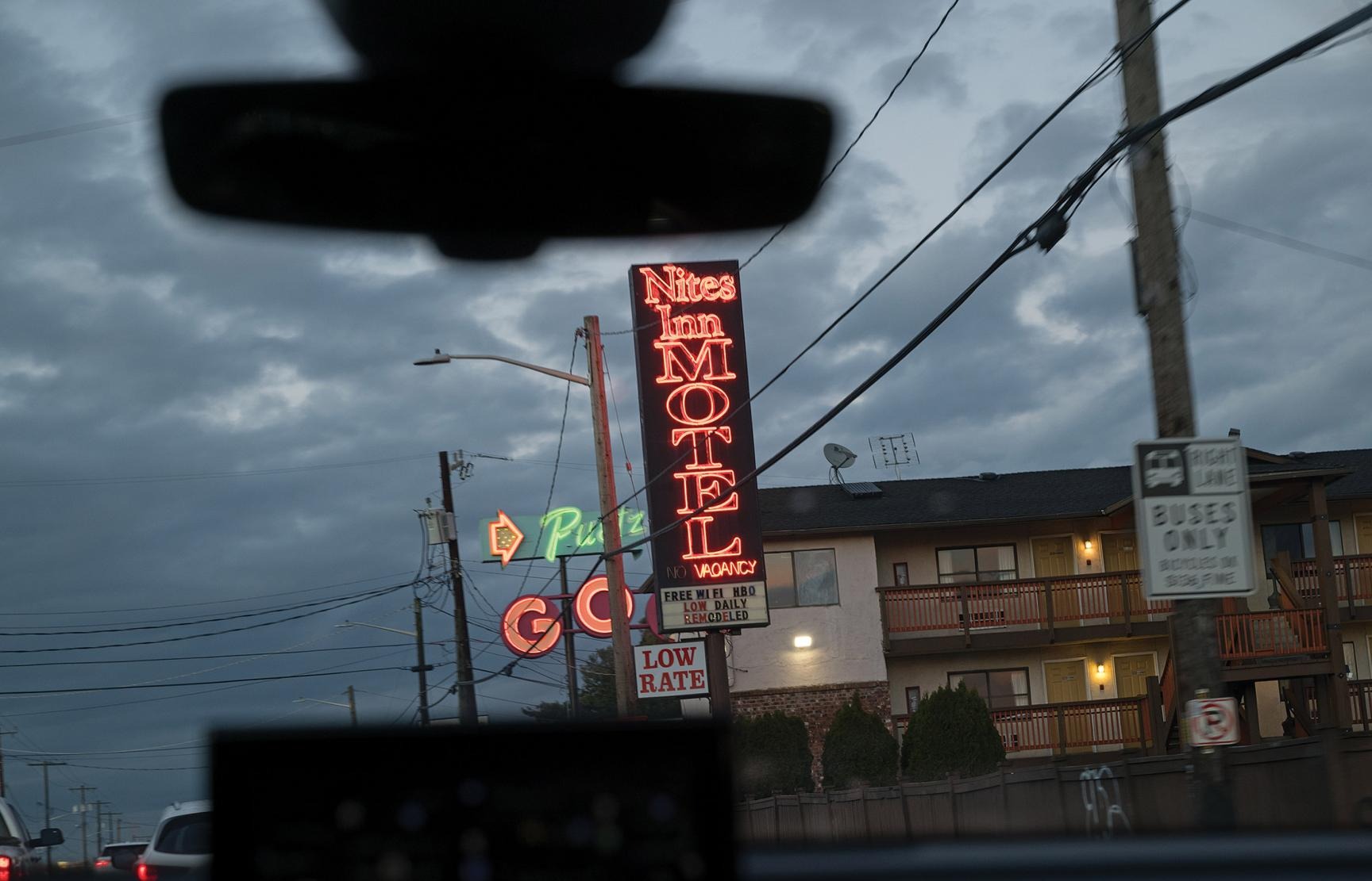 Neon sign reading "MOTEL" beside a lit building along a street with power lines under a cloudy evening sky