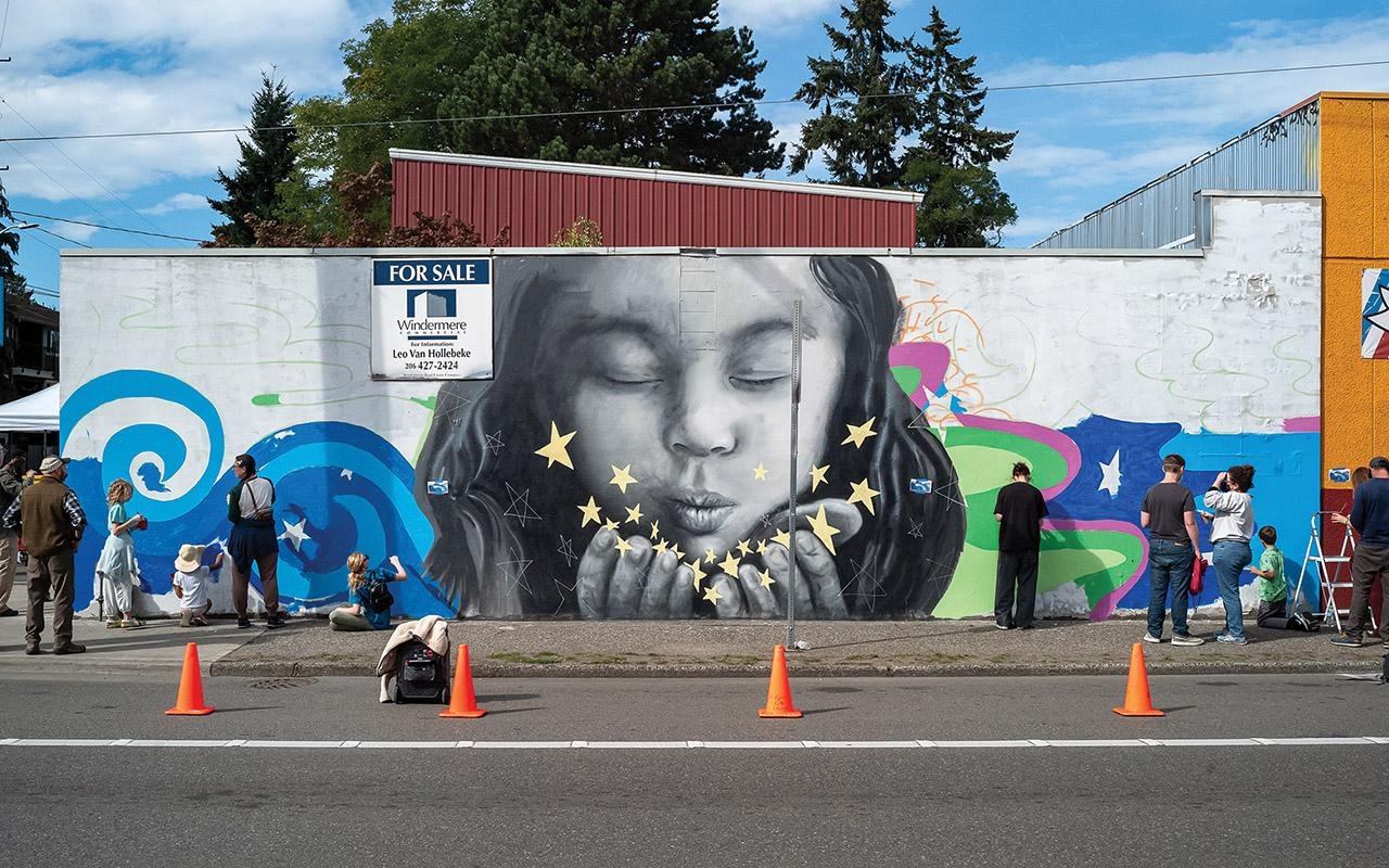Mural of a closed-eye face on a building wall, with people standing nearby and orange traffic cones along the street.