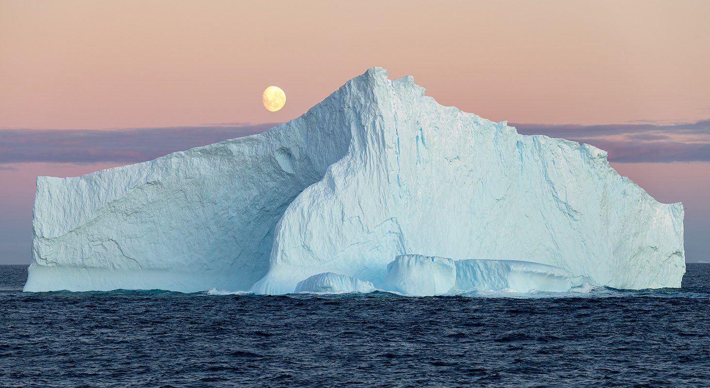 Un iceberg che galleggia in mare aperto con la luna visibile sopra l'orizzonte.