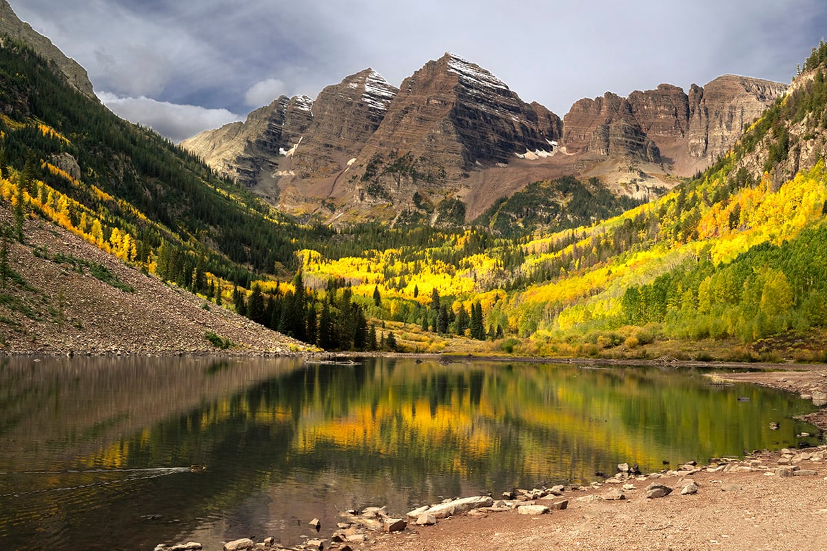 Cime montuose che si riflettono in un lago, con alberi gialli e verdi che costeggiano la riva.