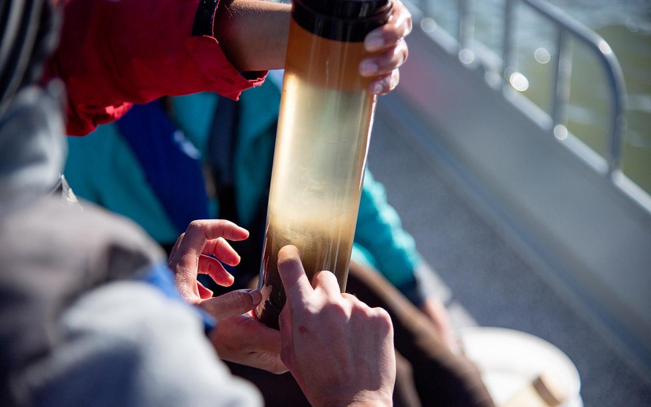 Hands holding a clear sampling tube containing water and sediment collected from a body of water.