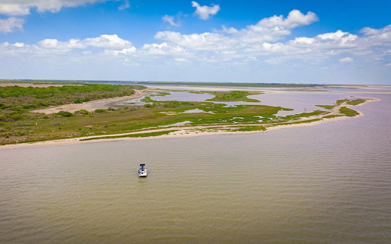 Aerial view of a small boat on a wide river near marshland and shoreline under a blue sky.