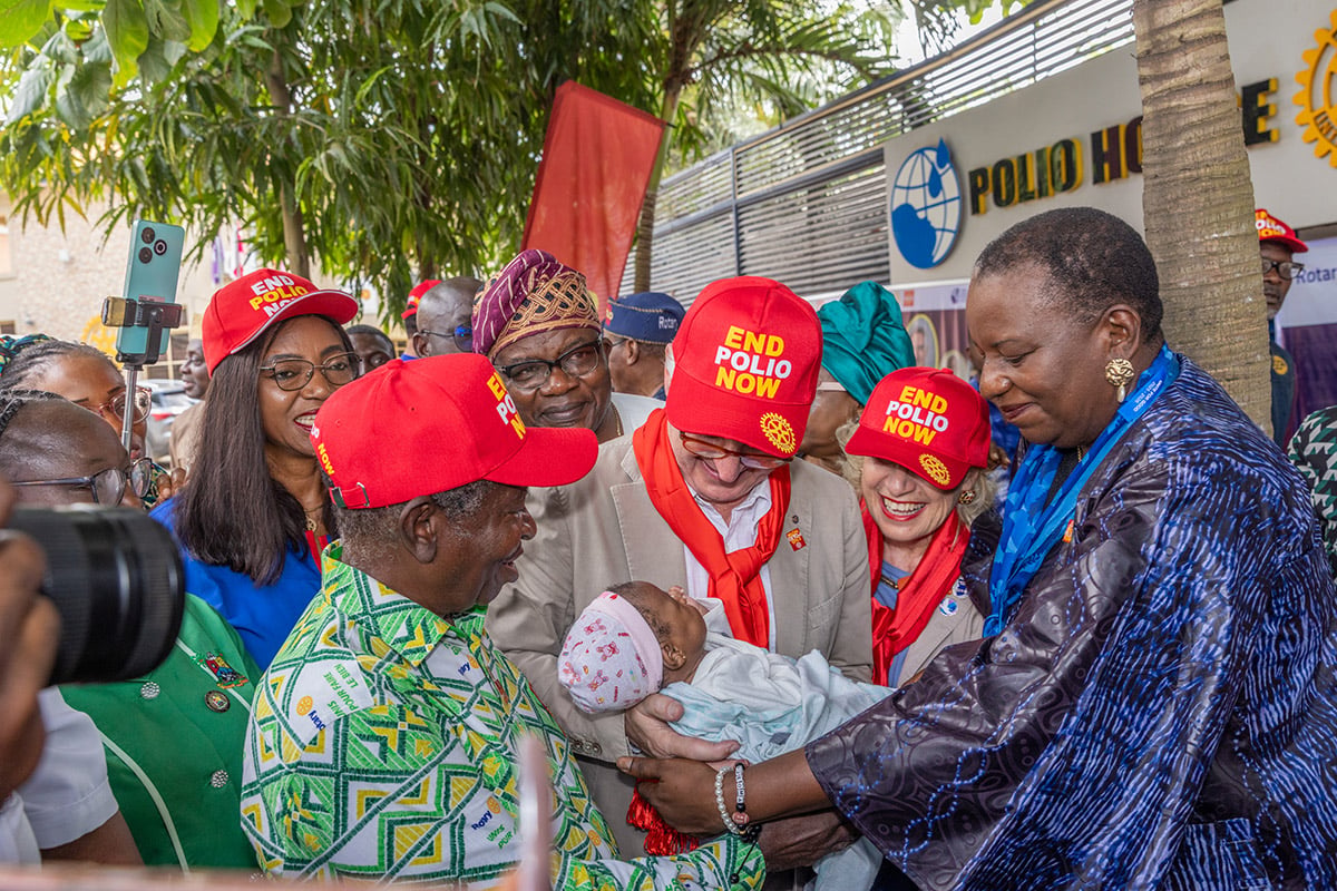 People wearing End Polio Now caps gather around and hold a baby outside a Polio House building.