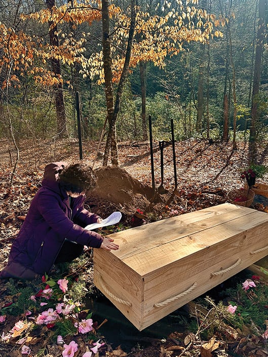 Person kneeling beside a wooden coffin at a graveside surrounded by flowers and shovels.