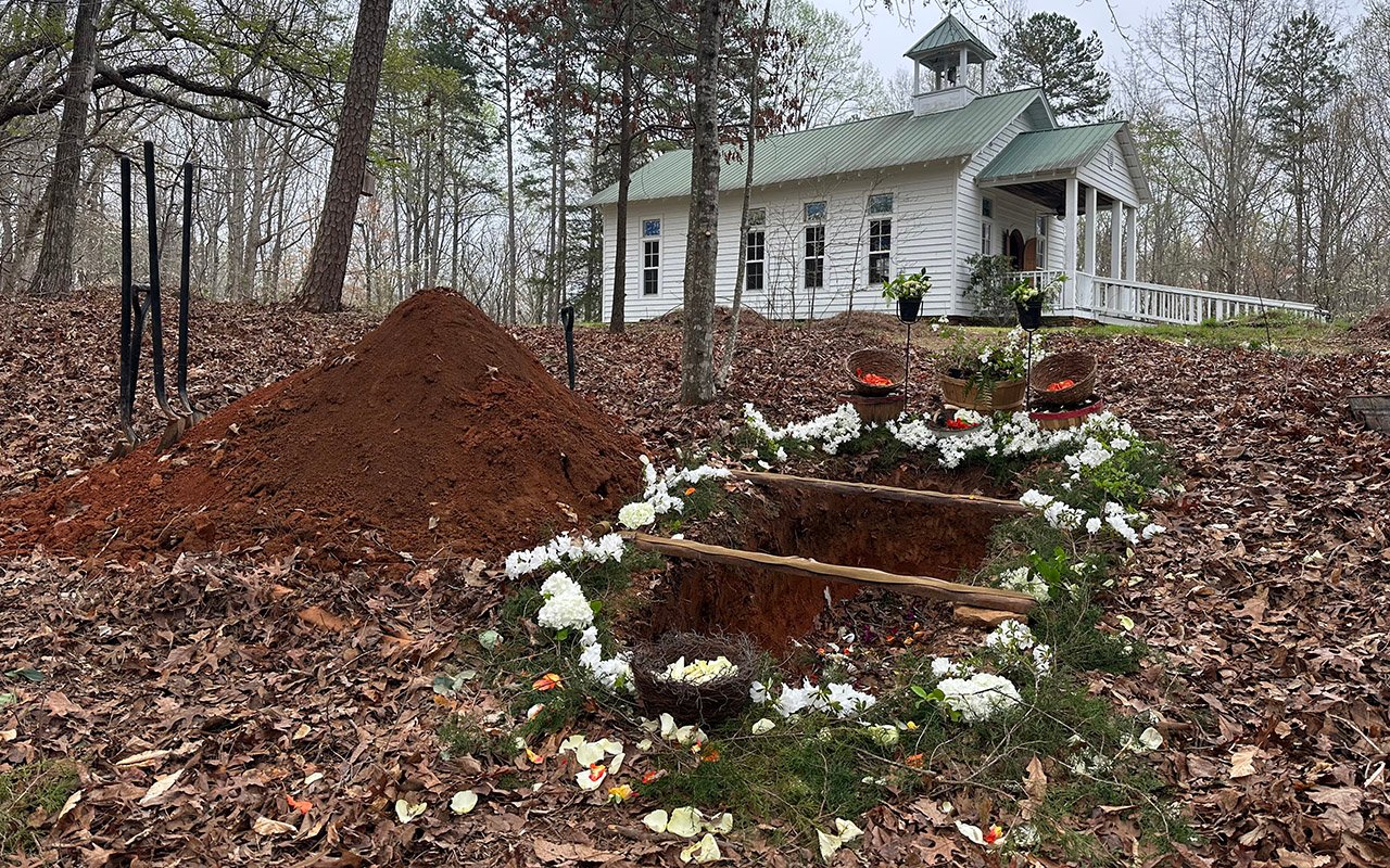 Open grave decorated with white flowers beside a church, with a mound of earth nearby.