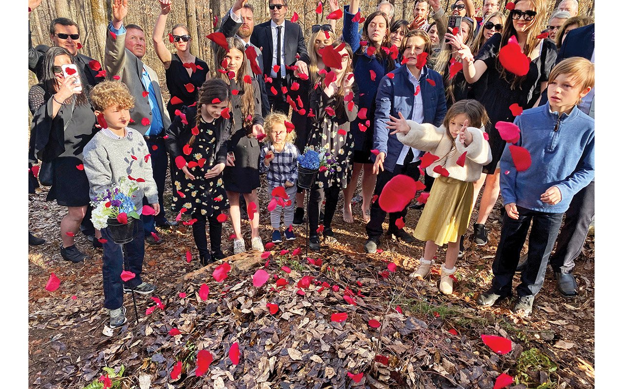 People gather around a gravesite and toss red flower petals over a mound of leaves.