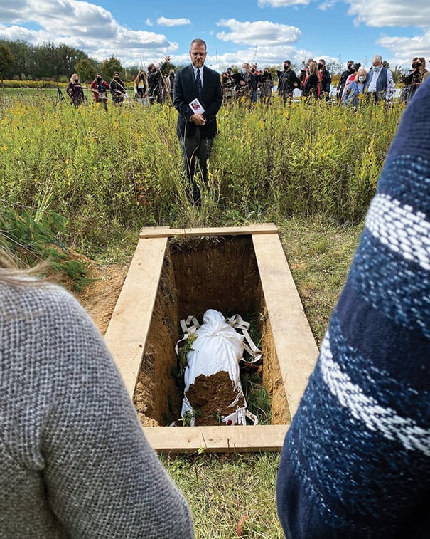 Open coffin in a gravesite as people gather in a field nearby.